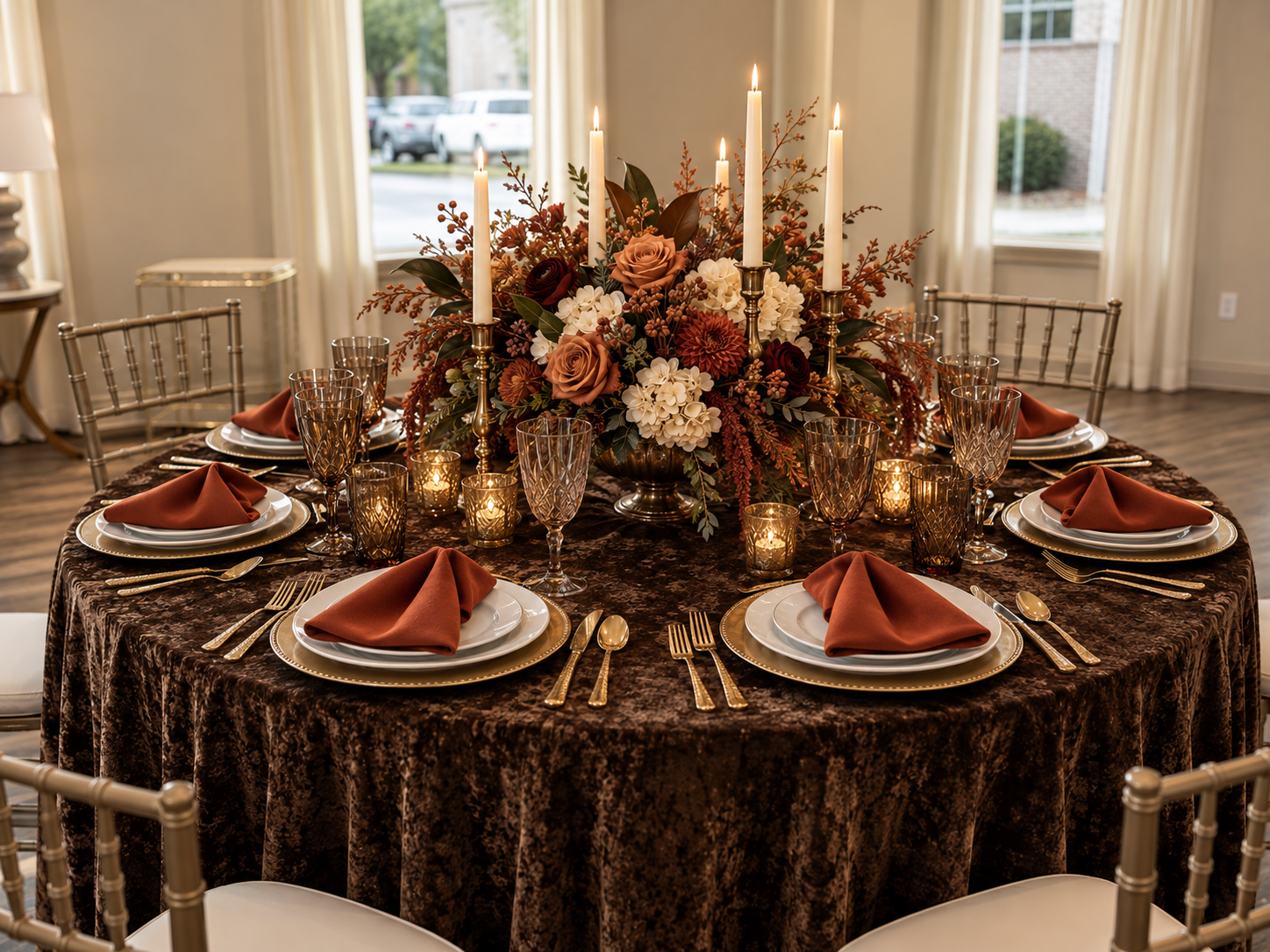 Chocolate & Cognac tablescape with a deep chocolate crushed velvet linen, cognac napkins, gold-rimmed chargers with white dinnerware, gold flatware, cognac cut-glass goblets, brass candlesticks with ivory tapers and glowing votives, and a lush centerpiece of caramel roses, burgundy blooms, ivory hydrangeas, and autumn foliage
