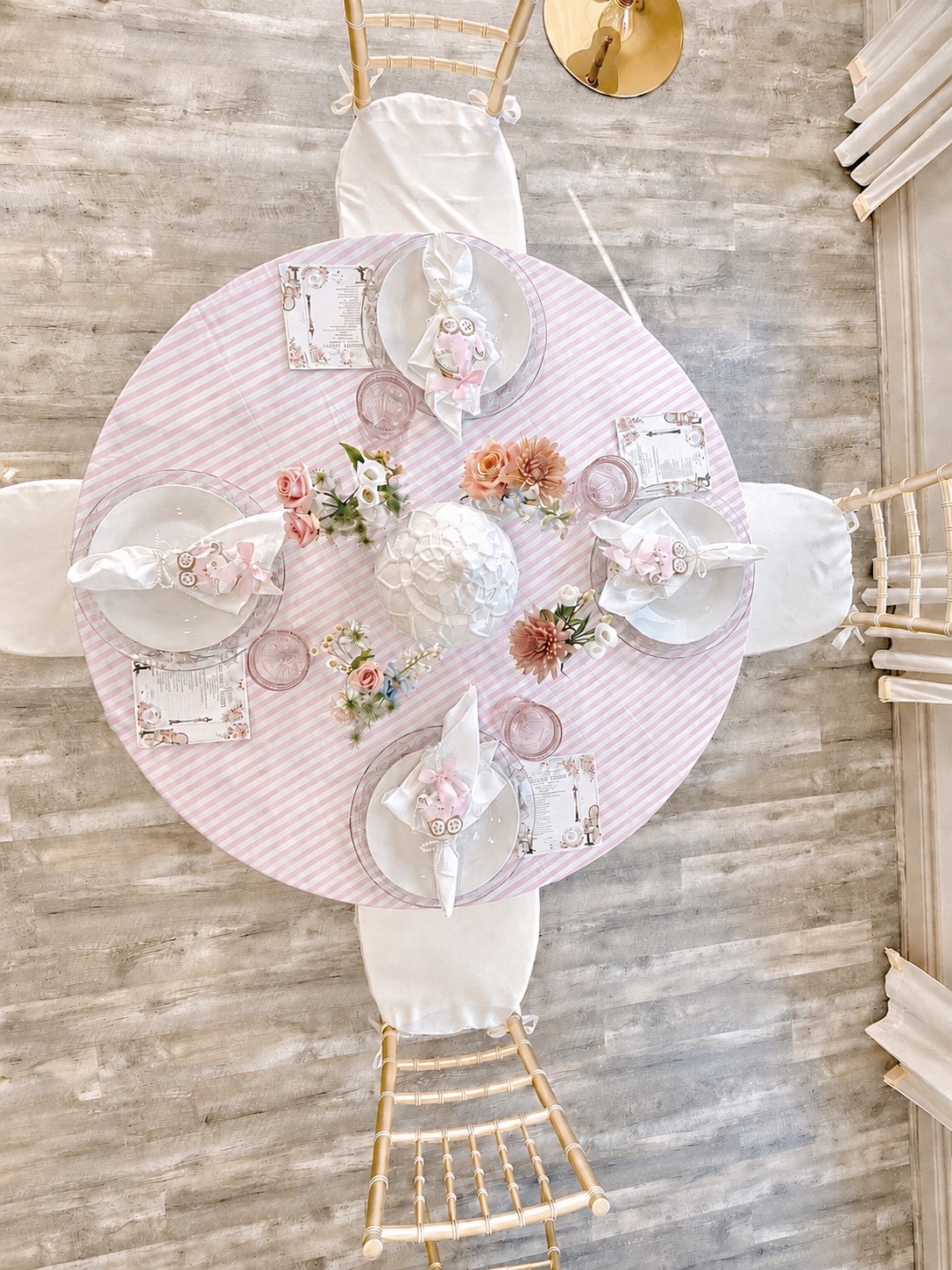 An overhead view of a Parisian-themed baby shower place setting with a pink striped linen, glass plates, dainty florals, and champagne gold chairs