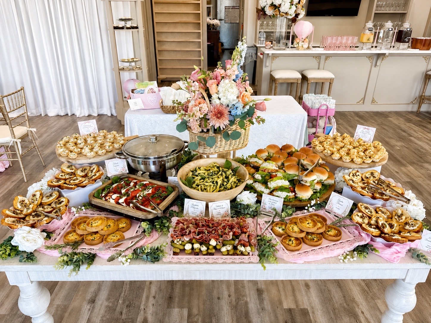 A Parisian-themed baby shower buffet at Magnolia Studio — sandwiches, quiche, bruschetta, and pasta salad styled with pink linens, lace trays, and a fresh floral basket